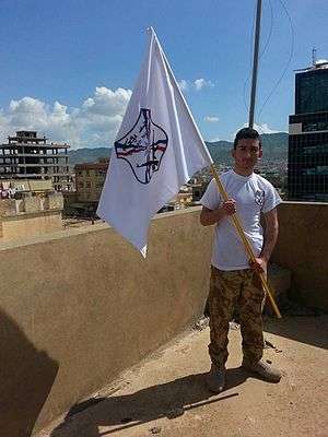 A Dwekh Nawsha fighter holding the militias flag and emblem, which features the Assyrian flag with two AK 47s crossed.