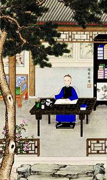Color painting of young man wearing a deep-blue robe and a black sleeveless jacket sitting at a table holding a brush. A book, a brush with a cup, an inkstone, and a bowl filled with water are also placed on the table, which is itself black with golden or yellowish flower and leaf patterns. The table is disposed in a courtyard. There is a large tree in the left foreground that runs from bottom to top. On the right we see part of a bookshelf with books on it. Part of a wide chair appears on the left. In the background is the entrance to a small building.