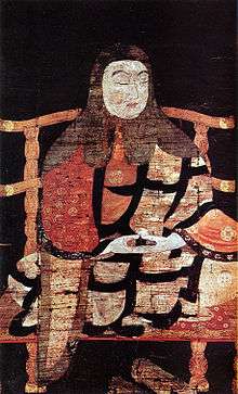 Portrait of a monk in three-quarter view seated cross-legged on a chair. His hands rest, palms up, on his lap in meditating pose.