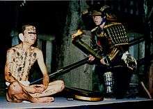 A man in black armor kneels beside a man clad only in a loincloth.  The second man has black symbols painted on his body and bandages over his ears.