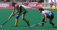 A female Australian player with blonde hair moves crouched over on the left of the screen while holding her hockey stick down low to move the ball. Behind her is a brown haired Argentinian player who is also crouched down and moving towards the Australian.