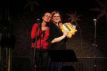 Photograph of the sisters almost back-to-back in front of a microphone stand, with Angela holding a cat-shaped electronic piano.