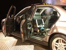 Side view of a vehicle with its doors open. Behind the left front seat can be seen a steering wheel and monitor.