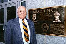 Elderly man in coat and tie standing next to a bronze dedication plaque located at the building entrance.