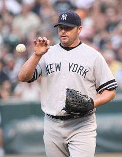 A man in a gray baseball uniform catches a baseball with his bare right hand. He is wearing a navy blue cap on his head with an interlocked "NY" and a black baseball glove on his left hand. His uniform reads "New York" across the chest.