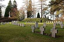 In the foreground, a collection of differently designed headstones in a green, mostly open, field surrounded by trees. In the background the German designed obelisk and British designed Cross of Sacrifice, each on raised hills, can be seen.