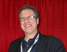 Smiling man with black shirt and glasses, in front of a curtain