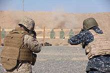 A Marine (left) and a sailor (right) both dressed in combat gear, fire at a target on a desert weapons range