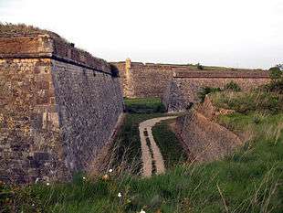 Photo shows a stone-faced fortress wall 20 to 30 feet high on the left. To the right is a stone wall 10 feet high. In between is a low area with a trail.