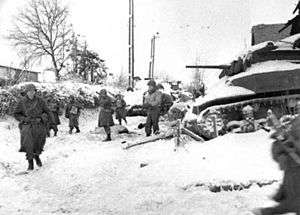 U.S. soldiers walking past a destroyed tank near St. Vith
