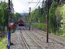 A double-track railway surrounded by trees runs down the middle of the image. On the left track is worn-looking red train. Above the tracks are overhead wires.