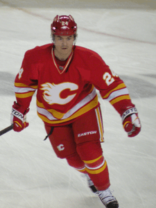 A hockey player in a red uniform with white and yellow stripes at the waist skates forward.  His uniform has a white, stylized C logo on the chest and the number 24 on his arms.