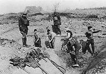 Five soldiers dig a trench, supervised by two standing above them.