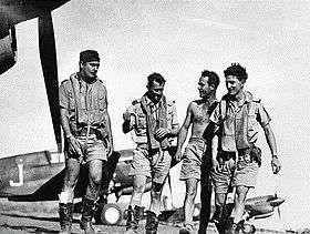 Four men in military flying gear walking among single-engined aircraft on an airfield