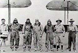 Seven men, four in flying suits, standing in front of the wings of two biplanes