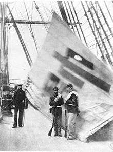 two Marines stand in front of and one sailor next to a white flag with a Chinese character, displayed from the rigging of a ship