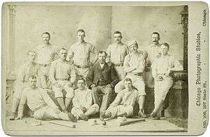 Twelve members of a baseball team are posing for a photograph, consisting of three rows; five men standing, four sitting in a chair or bench, and three sitting on the floor