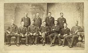 Baseball players are posing for a photograph, four men standing, seven men sitting on chairs.