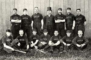 Two rows of men wearing old-style dark-colored baseball uniforms with "PHILA" on the chest; in the rear center stands a bearded man with a high dark top hat and a Victorian-era suit