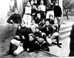 Football team photo outside a building; some players are standing, and others are sitting on the steps.