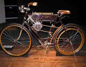 A pristine-looking, spindly turn-of-the-century style motorcycle with a chain and pedals like a bicycle, and a flame lantern for a headlight.  A leather belt delivers power from the engine to the rear wheel.
