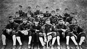 In this photograph of a baseball team, seventeen men wearing dark uniforms and caps are sitting on a raised parcel of dirt in three rows facing the camera.