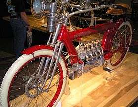 A bright red 1910 motorcycle with a flame headlamp, white rubber tires and a four-cylinder engine.