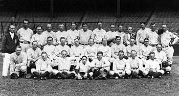 A posed team photo of a baseball team. They wear uniforms and are seated and standing in three rows, with a man in a jacket standing to the left.