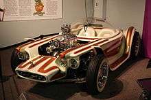 A white car with brown stripes, with open wheels and a clear bubble canopy over twin seats, and exposed, chromed engine with a blower.