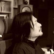 Sepia-toned square side profile image of Keiichi Oku. He has a light moustache and a thin beard along his jawline. He is looking toward the upper right corner of the image.