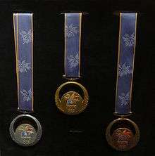 Three round medals with blue ribbons hanging in a display. The medals are silver, gold, and bronze from left to right.