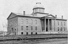 A black-and-white photograph of a long, Greek-revival building with tall pillars at the entrance and a great dome in the center.