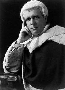 A man wearing a judge's wig and fur-tipped robes with his head supported on his right hand; his right elbow is on a pile of books