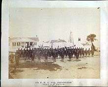 Union engineers lined up in sandy foreground of white houses, white lighthouse in the far background