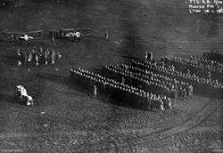 A large number of people standing in formation in a field. Two biplanes are visible at the upper left, with a smaller group of people standing in front of them.