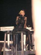 A blond woman stands on a steel tabletop. She wears a tight, body-hugging dress, accompanied by boots and gloves. She sings to a microphone in her left hand and her right hand is stretched out. The table is held by steel cables.