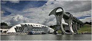 The Falkirk Wheel dominates the right of the picture with the tourist shop and restaurant dwarfed by the Wheel on the left, the bottom shows the marina with a boat in the centre