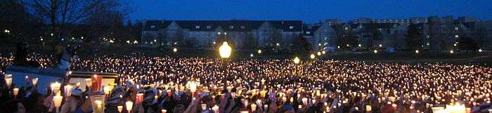 A sea of candles shining in the darkness of the Drillfield with campus buildings on the opposite side.