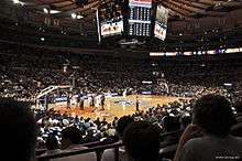 A basketball arena viewed from far away in the stands during a game. The overhead scoreboard and entire court are in view while a player at the near free throw line is shooting free throws.