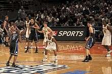 Two opposing basketball teams on a basketball court with a team in white defending the basket behind them. One team is in white uniforms with the word Michigan on the front and names on the back and the other is dark blue with the word Duke on the front and names on the back.