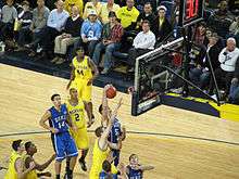Two opposing basketball teams on a basketball court fight for a rebound. One team is in maize uniforms with the word Michigan on the front and names on the back and the other is light blue with the word Duke on the front and names on the back.