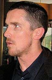 Profile shot of a man looking to his left. He has a goatee and stubble, and is wearing a black dress shirt with collar and a black blazer.