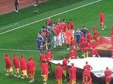 A team in blue shakes hands with a team in red. In the foreground are people wearing red shirts and gold shorts assisting with the presentation of the game.
