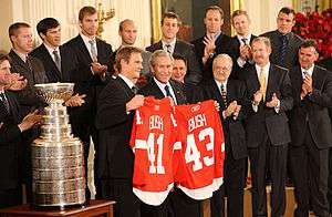 In the front row, Nicklas Lidstrom holds a Red Wings jersey with the last name "BUSH" and the number "41" while George W. Bush holds a Red Wings jersey with the last name "BUSH" and the number "43".  To Lidstrom's right is the Stanley Cup.  2nd row, left to right: Mike Babcock, Ken Holland, Jim Devellano, Jim Nill, unknown man.  Third row, left to right: Kris Draper, Pavel Datsyuk, Mikael Samuelsson, Brian Rafalski, Derek Meech, Kirk Maltby, Chris Osgood, Aaron Downey.