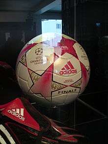 A football in a perspex cabinet. The ball is predominantly white, patterned with stars in red with gold detailing. In the foreground is a black football boot with red and white trim.