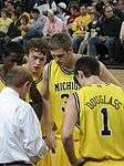 The basketball players in maize uniforms are huddled around a man in a white shirt.