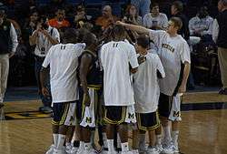 A team of basketball players are huddled together on a basketball court. The entire team is reaching to the center with one arm so that their hands touch.