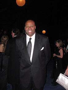 Craig Robinson is smiling for the camera while attending a very formal ball. He is wearing a dark black suit that has visible grey-colored lines running vertically through it. His white undershirt is complemented by a shiny, solid black tie.
