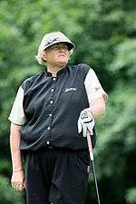 A blonde-haired woman in a black vest, pants, and hat and a white golf undershirt is holding a golf club