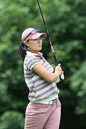 A dark-haired woman is wearing lavendar colored pants with a shirt that has colorful geometrical shapes on it with a lavendar hat and sunglasses on the bill of the cap and a graphite shafted golf club after a golf swing is up in the air